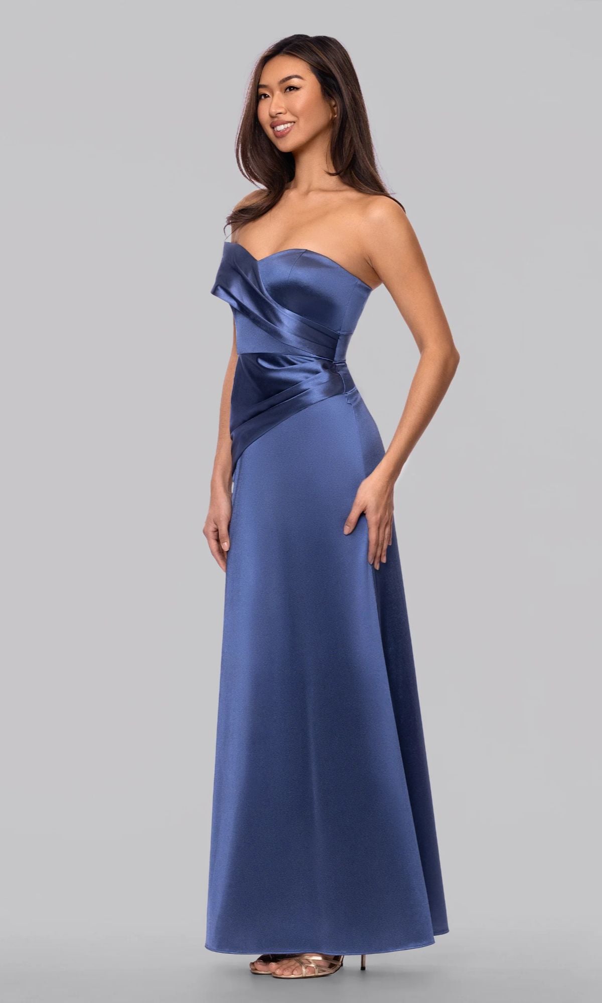 A woman in the Blondie Nites 6164BN long prom dress—floor-length, off-the-shoulder slate blue satin with a fitted bodice and asymmetrical detail—stands against a plain gray background.